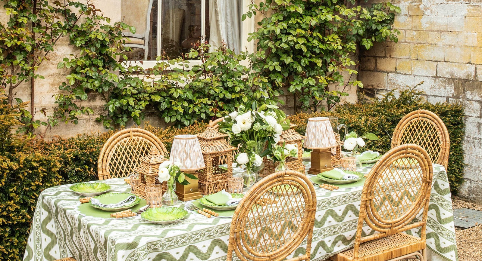 Outdoor dining setup with wicker chairs and a patterned tablecloth in a garden setting.